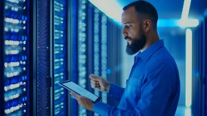 IT technician inspecting server cables and equipment in a data center, using a tablet for diagnostics and system monitoring.